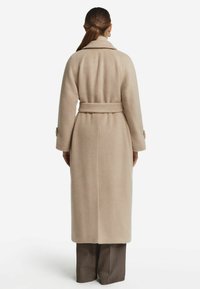 Kvinde med samlet hår iført en lang beige belted frakke og brune brede bukser, vendt væk mod en ensfarvet hvid baggrund.