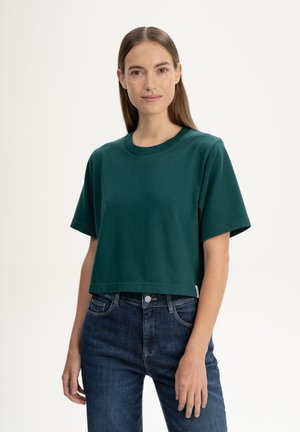 Vrouw met stijlbruin haar die een donkergroen cropped T-shirt en donkerblauwe jeans draagt, tegen een effen witte achtergrond.