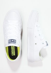 Zapatillas de lona blancas con suelas de goma, puntera redonda y diseño clásico con cordones. Presentan una plantilla negra y verde y un logotipo en la lengüeta.