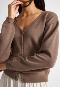 Cardigan en tricot marron avec un col en V, fermeture à boutons à l'avant et des poignets et un ourlet côtelés. Texture douce, coupe décontractée et éclat subtil.