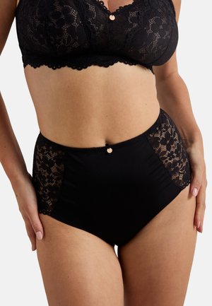 Femme portant un soutien-gorge en dentelle noire et une culotte taille haute, les mains posées sur les hanches, devant un fond clair uni.