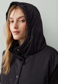 Schwarze Kapuzenjacke mit glatter, gl änzender Textur, Druckknöpfen und Stehkragen. Das Modell hat lange Haare und eine klare Haut.