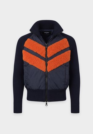 Homme portant une casquette marron, une veste noire et orange, un t-shirt blanc avec un graphisme, un jean bleu usé avec une ceinture marron et des bottes marron, debout sur fond blanc.