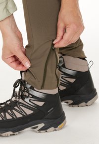 Mörkgröna utomhusbyxor med halvvippa vid ankeln, ihop med svarta och grå vandringsskor som har texturerade detaljer och en robust sula.