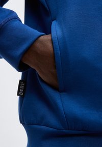 Σκούρο δέρμα χεριού εισάγεται στην μπροστινή τσέπη μιας εφαρμοστής μπλε φούτερ με μαύρη ετικέτα που φέρει λευκούς ιαπωνικούς χαρακτήρες.