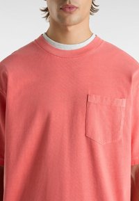 Personne portant un t-shirt corail rose à col rond avec poche poitrine sur un sous-t-shirt gris clair à col rond, montré du cou à la taille.