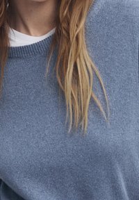 Blue knitted sweater with a round neckline and ribbed cuffs, featuring a soft texture. Below, light brown hair with natural waves.