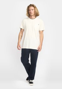 Jonge man met schouderlang blond haar, gekleed in een wit T-shirt met een borstzakje, marineblauwe broek, zwarte sneakers en een kralenketting, staand en lopend.