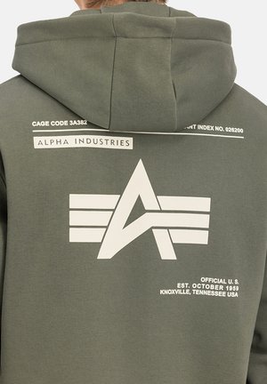 Olivegrüner Hoodie mit einem großen weißen Logo, das ein „A“ und Flügeldesign zeigt, begleitet von Text, der die Marke und Herkunft beschreibt.