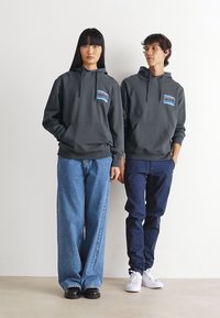 Twee mensen in donkere hoodies met frontzakken en grafische patches, staan zij aan zij tegen een effen witte achtergrond.