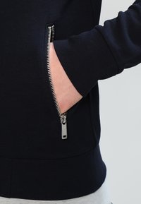 Námornícky sveter s kapsou na zip, která má stříbrný zip a hladkou texturu. Detailní pohled na kapsu.