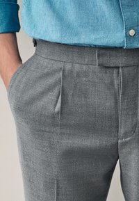 Pantalon gris sur mesure en tissu texturé, avec plis avant et poches latérales. Accompagné d'une chemise en denim bleu.