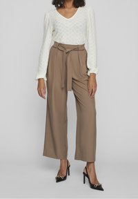 Haut crème texturé à manches longues avec décolleté en V, associé à un pantalon brun taille haute et large, et des talons noirs pointus avec des accents dorés.