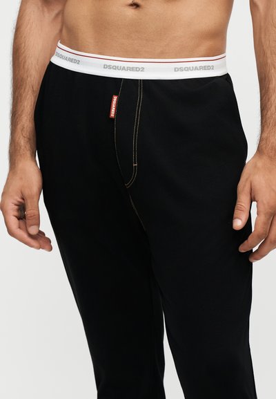 Homme portant un pantalon noir avec une ceinture élastique blanche visible marquée DSQUARED2 et une étiquette rouge DSQUARED2 près de la couture sur la zone avant de la braguette.
