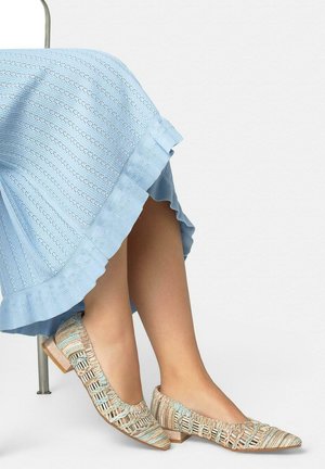 Gekruiste benen met beige en turquoise geweven puntige platte schoenen, met een lichtblauwe gebreide rok over de knieën gedrapeerd tegen een effen achtergrond.