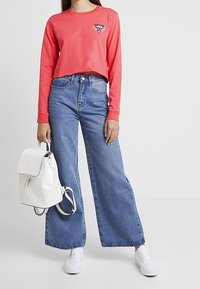 Korallefarbenes Langarm-Cropped-T-Shirt mit Logo, kombiniert mit weit geschnittenen blauen Jeans. Ein weißer Rucksack und Sneaker runden das Outfit ab.