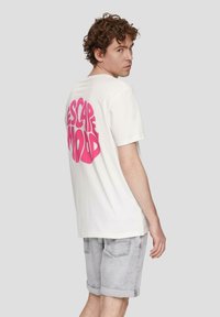 Weißes Baumwoll-T-Shirt mit kurzen Ärmeln, auf der Rückseite mit großem pinken "ESCAPE MOOD"-Schriftzug. Das Modell kombinierte es mit hellgrauen Jeansshorts.