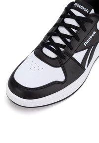 Adidași negri și albi cu o parte superioară din piele, vârf rotund și perforații. Prezintă șireturi negre și logo-ul Reebok pe lateral.