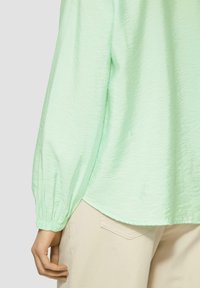 Hellgrünes, strukturiertes Langarmshirt mit gerafften Manschetten, kombiniert mit cremefarbenen Hosen. Das Shirt hat eine lockere Passform und einen abgerundeten Saum.