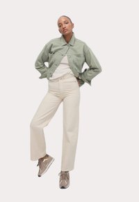 Giacca di denim verde chiaro, camicia bianca e pantaloni ampi color crema; modella che indossa sneakers grigie, in una posa rilassata.