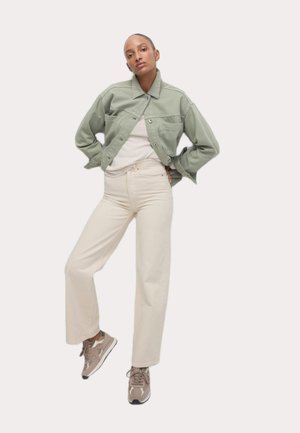 Veste en denim vert clair, chemise blanche et pantalon large crème ; modèle portant des baskets grises, adoptant une pose décontractée.