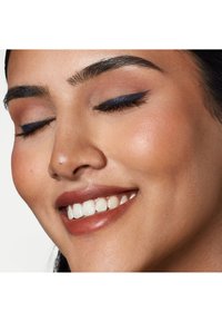 Närbild av ett leende ansikte med en jämn, varm hudton. Drag som framhävs inkluderar kraftig eyeliner, naturliga läppar och ett subtilt rouge på kinderna.