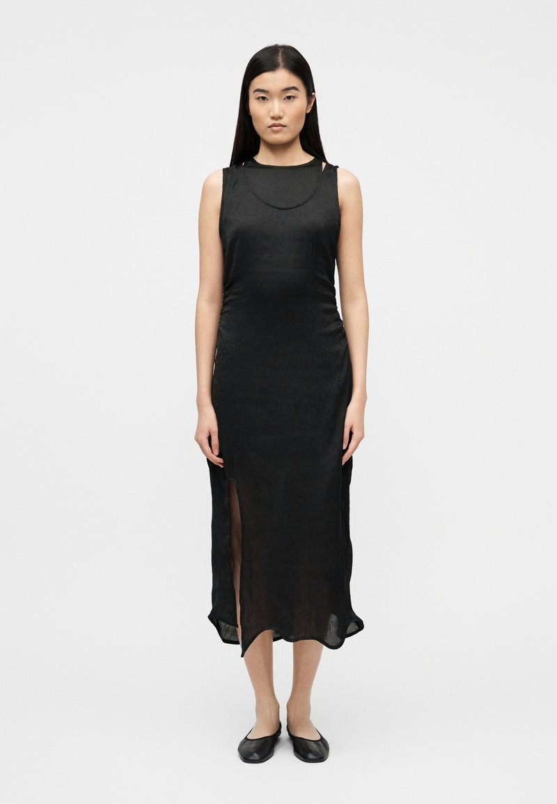 Γυναίκα που στέκεται με το πρόσωπο προς τα εμπρός, φοράει μαύρο αμάνικο μεσαίο φόρεμα με σχίσιμο μπροστά και μαύρα επίπεδα παπούτσια σε λευκό φόντο.