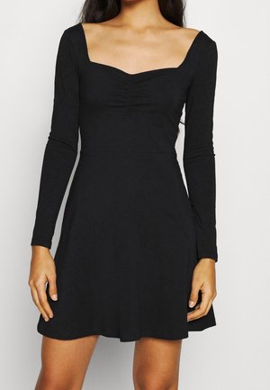 Kobieta ubrana w czarną, długorękawową sukienkę w kształcie litery A z marszczonym dekoltem typu sweetheart na jednolitym tle.