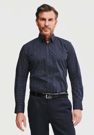 Granatowa koszula zapinana na guziki z białymi kropkami, charakteryzująca się gładką teksturą materiału i dopasowanym krojem. Czarny pasek dopełnia całość.