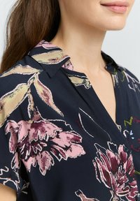Camicia con stampa floreale su base blu scuro, caratterizzata da fiori rosa e gialli. Tessuto morbido e texture, con colletto classico e dettaglio con un solo bottone.