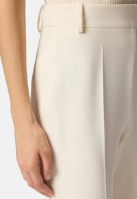 Pantaloni bianchi su misura con una texture liscia, caratterizzati da pieghe, vita aderente e dettagli con passanti per cintura. Mano appoggiata sul tessuto.