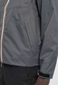 Grå lett jakke med hel glidelås, kontrastfargede detaljer og strukturert stoff. Inkluderer elastiske mansjetter og sidelommer.