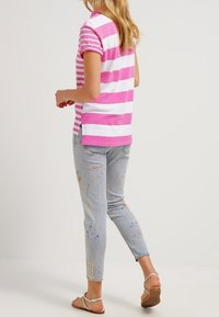 Mujer vestida con una camisa de polo a rayas rosa y blanca, jeans ligeros salpicados de pintura azul y sandalias blancas con tiras, de pie sobre un fondo liso.