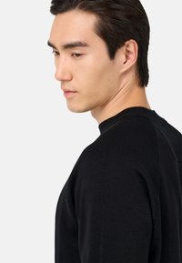 Juodas megztinis su apvalia iškirpte ir raukšlėmis ant rankovių. Audinys turi lygų paviršių ir prigludusią siluetą, rodomas šoninėje pusėje.