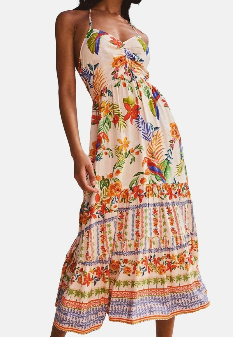 Femme portant une robe midi sans manches avec des imprimés floraux tropicaux colorés et des feuilles sur un fond clair.