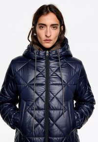 Námornícky prešívaný puffer kabát s kapucňou, s obdĺžnikovým uzatváraním a sťahovacími šnúrkami, vyrobený z lesklého materiálu s diamantovým vzorom a bočnými vreckami.