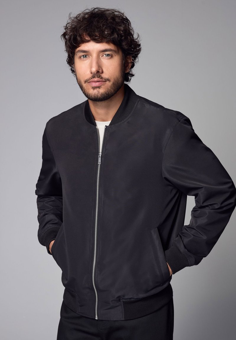 Giacca bomber nera con zip frontale, colletto e polsini a coste, texture liscia e tasche laterali. Indossata sopra una camicia chiara.
