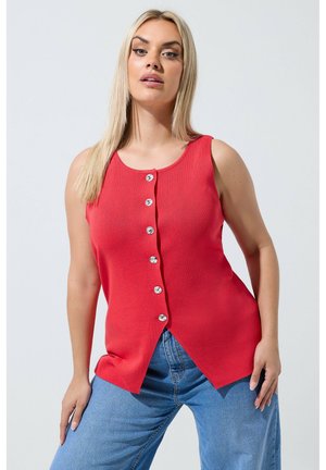 Ung kvinde med langt blondt hår iført en ærmeløs rød knapbluse og blå jeans, stående mod en ensartet lys baggrund.
