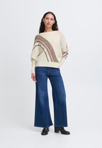 Maglione color crema con motivi a righe marroni, vestibilità morbida e maniche larghe. Abbinato a jeans a gamba larga blu scuro e stivali neri alla caviglia.