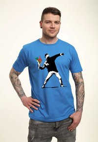 Modrý tričko s krátkým rukávem s černobílou grafikou osoby, která hází květiny, se červenými a zelenými akcenty. Uvolněný střih.