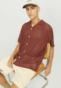 Camisa de manga corta de punto abierto en color óxido, con cuello y botones frontales, combinada con pantalones cortos beige claros, contra un fondo neutro.