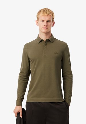 Olijfgroene polo met lange mouwen van katoen, met een drieknopenlijst en geborduurd logo op de linker borst. Model houdt een jas vast.