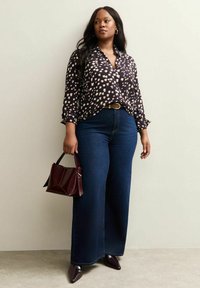 Granatowa koszula z długim rękawem w biało-beżowy wzór, zestawiona z niebieskimi dżinsami z wysokim stanem i szerokimi nogawkami. Trzyma małą bordową torebkę.