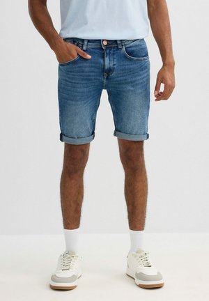 Hombre con pantalones cortos de mezclilla azules remangados, calcetines blancos y zapatillas blancas, de pie frente a un fondo claro y liso.