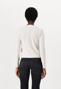 Personne aux cheveux courts et bouclés portant un pull blanc à manches longues et un jean noir ajusté, debout de dos devant un fond uni.