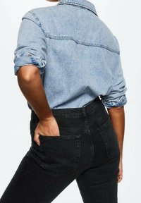 Chemise en denim bleu clair aux manches retroussées, associée à un jean noir taille haute avec poche arrière et légère élasticité pour le confort.