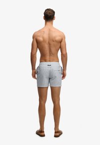 Homme musclé debout pieds nus, dos tourné vers la caméra, portant un short de bain bleu clair à carreaux et des sandales marron sur fond blanc.