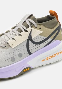 Chaussure de trail Nike ZoomX en gris clair et vert olive, avec un dessus texturé, une semelle intermédiaire épaisse violette et des accents orange.