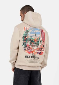 Hombre vistiendo una sudadera beige con un gráfico colorido de un balcón parisino, dos sillas, plantas y vista de la Torre Eiffel en la parte posterior.