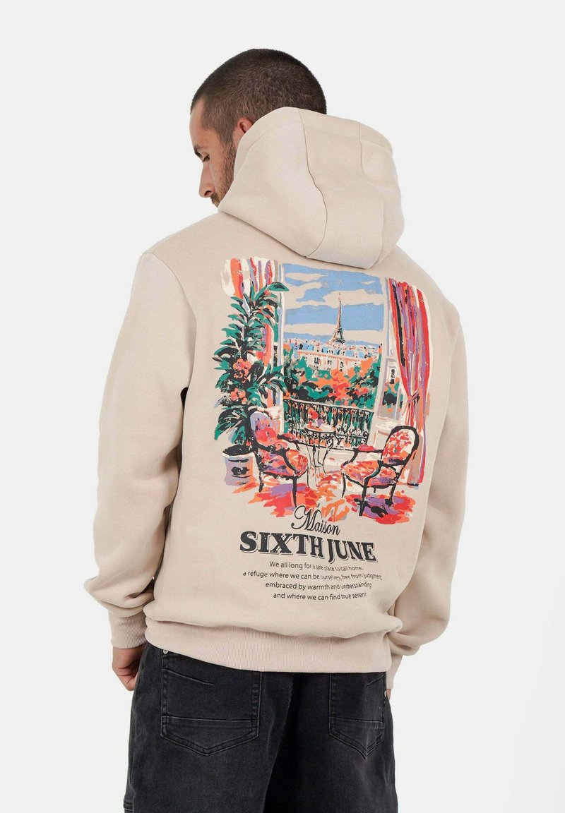 Hombre vistiendo una sudadera beige con un gráfico colorido de un balcón parisino, dos sillas, plantas y vista de la Torre Eiffel en la parte posterior.